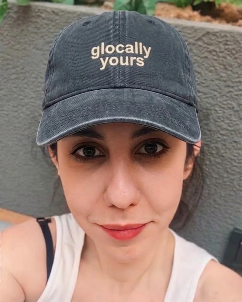 Portrait of Işıl Öztemel wearing a dark cap embroidered with “glocally yours”